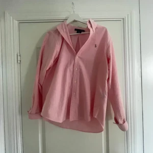 Rosa skjorta från Ralph Lauren - Snygg rosa skjorta från Ralph Lauren med klassisk krage och knappar framtill. Liten broderad logga på bröstet. Skjortan har lång ärm och är gjord i mjuk bomull. Perfekt för dig som gillar stilrena plagg med en touch av färg.ny pris 1600kr men jag säljer för endast 499