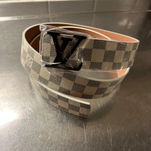  rutig beige bälte - Snyggt bälte från  med klassiskt rutigt mönster i beige och grått. Bältet har en stor LV-logga i svart metall som spänne och är tillverkat i skinn. Perfekt för dig som vill ha en exklusiv och trendig accessoar med lyxig känsla.