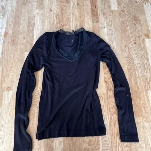 Svart långärmad tröja med spets - Svart långärmad tröja med v-ringad hals och snygga spetsdetaljer runt halsringningen. Mjuk och tunn, perfekt för lager på lager. Tröjan har en feminin touch med spetsen och sitter snyggt på kroppen. Passar dig som gillar stilrena och enkla plagg med en twist.