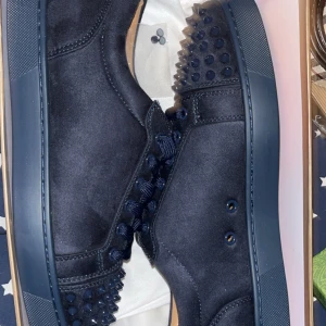 Mörkblå Christian Louboutin sneakers - Säljer ett par mörkblå sneakers från Christian Louboutin med ikonisk röd sula och coola nitar framtill. Skorna är gjorda i mocka och har snörning med matchande mörkblå skosnören. Perfekt för dig som vill sticka ut med lyxig vibe och edgy detaljer.