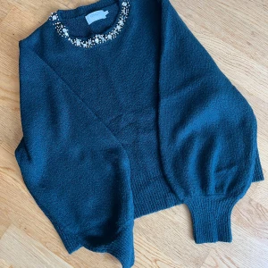 Mörkgrön stickad tröja med pärlor ONLY - Snygg mörkgrön stickad tröja från ONLY med pärl- och stenbroderi runt halsen. Tröjan har långärmade ballongärmar och ribbade muddar vid ärmslut och nederkant. Strl XXL.