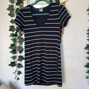 Svartvit randig kortklänning Nanso - Svart kortklänning från Nanso i storlek S med vita horisontella ränder. Klänningen har v-ringad hals och korta ärmar, vilket ger en chill och enkel look. Materialet känns mjukt och lätt, perfekt för varma dagar. Passformen är normal och klänningen är superenkel att styla.