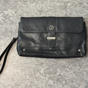 Armani Jeans svart kuvertväska - Stilren svart herrkuvertväska från Armani Jeans i mjukt skinn. Väskan har ett praktiskt handledsband, silverfärgade metalldetaljer och flera fack med dragkedja. Perfekt storlek för det viktigaste och snygg AJ-logga framtill. Ny, aldrig använd.