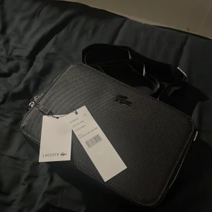 Svart axelväska från Lacoste - Snygg svart axelväska från Lacoste med stilren design och präglad yta. Väskan har justerbar axelrem, silverfärgad dragkedja och ett diskret Lacoste-logo framtill. Materialet är syntet med struktur, och insidan har en praktisk ficka med dragkedja.