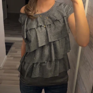 Grå glittrig volangblus  - Säljer en vintage grå glittrig blus med flera lager volanger framtill och baktill. Toppen är i bra skick och inga skador eller fläckar! Skriv om det finns frågor💞
