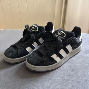 Adidas Campus svarta sneakers strl 42 - Svarta Adidas Campus sneakers med klassiska vita ränder och logga på plösen och hälen. Skorna har snörning, rund tå och platt grå sula. Ovandelen är i mocka och detaljerna i syntet. Perfekt för dig som gillar retrostil och streetwear. Endast använda fåtal gånger och därför inga tecken på användning. Kom helt enkelt inte till användning. MYCKET fint skick!!🤩🥰