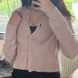 Ljusrosa stickad kofta från Lindex - Superfin ljusrosa stickad kofta från Lindex i storlek S. Koftan har stora matchande knappar framtill, v-ringad hals och ribbade muddar. Mjuk och fluffig känsla, perfekt att slänga på sig när det är lite kyligt ute. Passar dig som gillar en söt och chill stil.