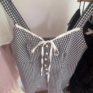 Svartvit rutig topp med pärlknappar - Supersöt svartvit rutig topp med breda axelband och vit snörning framtill. Klänningen har pärlknappar längs bröstet och en vit kant upptill som ger en extra touch. Perfekt för dig som gillar classyvibbar och vill sticka ut med detaljer.