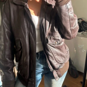 Brun skinnjacka med fickor - Säljer en brun skinnjacka med coola detaljer som stora fickor med knappar och ribbade muddar vid ärmslut och nederkant. Jackan har krage och dragkedja framtill, perfekt för dig som gillar vintagevibbar och vill ha en unik look.