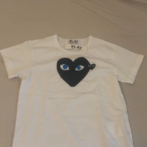  Comme des Garçons T-shirt  - Snygg vit t-shirt från Comme des Garçons Play i storlek S. Framsidan har ett stort svart hjärta med blå ögon, en ikonisk detalj från Play-kollektionen. Klassisk rund hals och korta ärmar. Tillverkad i mjuk bomull för skön passform. Passar som en xs