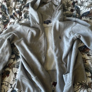 Grå hoodie från Polo Ralph Lauren - Säljer en grå hoodie från Polo Ralph Lauren med klassisk huva och dragkedja framtill. Tröjan har långa ärmar, två fickor och den ikoniska lilla Polo-loggan broderad på bröstet. Perfekt för chill dagar eller när du vill vara extra bekväm.