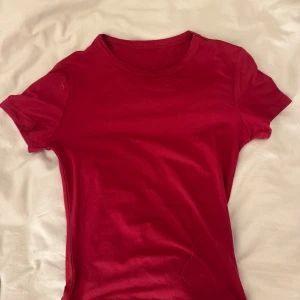 Röd basic t-shirt med rund hals - En enkel röd t-shirt med rund hals och korta ärmar. Modellen är klassisk och passar perfekt till jeans eller shorts. Materialet känns mjukt och skönt mot huden, troligtvis bomull. Perfekt för dig som gillar stilrena plagg i starka färger.