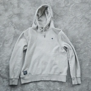 Ljusgrå hoodie från Morris Stockholm - Snygg ljusgrå hoodie från Morris Stockholm med broderad logga på bröstet och patch vid nederkanten. Tröjan har huva med snörning och långa ärmar, perfekt för chill dagar. Tillverkad i mjuk bomull som känns skön mot huden.