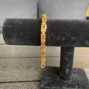 Guldigt länkarmband med chunky design - Snyggt guldigt armband med chunky länkar som ger en cool och modern vibe. Länkarna är rektangulära och har en blank finish som verkligen poppar. Perfekt för dig som vill ha ett statement piece till din outfit. Lätt att matcha med andra smycken och accessoarer.