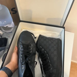 Svarta höga sneakers från Gucci - Säljer ett par svarta höga sneakers från Gucci med klassiskt GG-mönster på sidan. Skorna har snörning, rund tå och platt sula. Materialet är en mix av textil och skinn, med metallöglor för snörningen. Perfekt för dig som vill ha en lyxig och stilren look.