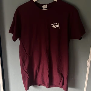 Vinröd Stüssy t-shirt med logga - Snygg vinröd t-shirt från Stüssy med vit logga tryckt på bröstet och i större format på ryggen. Klassisk passform och rund hals. Perfekt för dig som gillar streetwear och vill sticka ut med en fet logga. Materialet är mjuk bomull.