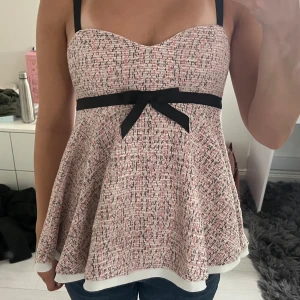 Mönstrad topp från ZARA med rosett och svarta band - Supersöt rutigaktig topp från Zara med svarta axelband och en svart rosett framtill. Blusen har en utsvängd form/volanger och är sydd i ett strukturerat tyg med rosa, svart och vit ton. Den är helt ny med prislapp och jag säljer då den var för tajt på mig!🥰 Storlek S men är liten i storleken.