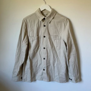 Beige overshirt från ARKET i bomull - Snygg beige overshirt från ARKET i ekologisk bomullstwill. Klassisk krage, svarta knappar framtill och på ärmslut, samt två stora bröstfickor. Perfekt lager-på-lager-plagg med clean look och normal passform. 1A21