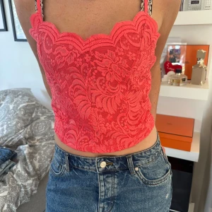 Rosa spetsig croppad linnetopp - Säljer en sjukt snygg croppad linnetopp i stark rosa med spets och blommigt mönster. Toppen har tunna justerbara axelband och en scallop-kant upptill. Perfekt att styla med jeans eller kjol för en cool look.