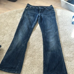Rock & Republic blå bootcut jeans - Snygga blå jeans från Rock & Republic med bootcut passform och låg midja. Baksidan har stora fickor med glittriga broderade loggor i silver. Jeansen har coola detaljer som blingade knappar och klassisk femficksdesign. Perfekt för dig som gillar statement-plagg.