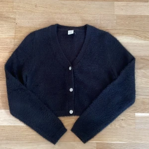 Svart fluffig kofta från Lindex 146/152 - Mysig svart kofta från Lindex med fluffig känsla och croppad passform. Koftan har långa ärmar och tre glittriga knappar framtill.