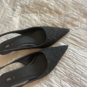 Svarta spetsiga pumps från Zara - Säljer ett par svarta pumps från Zara med spetsig tå och låg klack. Skorna har en glittrig textil på ovansidan och en öppen häl med smal rem. 