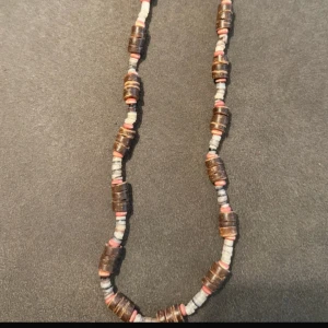 Brunt och grått pärlhalsband - Unikt halsband med runda pärlor i brunt, grått och inslag av rosa. Pärlorna är platta och staplade, vilket ger ett coolt och avslappnat uttryck. Perfekt accessoar för att lyfta din outfit med lite boho-vibe.
