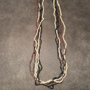 Flerfärgat pärlhalsband i bohostil - Säljer ett långt halsband med flera rader små pärlor i svart, brunt och beige. Halsbandet har en avslappnad boho-vibe och passar perfekt för dig som gillar naturliga färger och unika accessoarer. Pärlorna är oregelbundna och ger ett coolt, handgjort intryck. Står ej för frakt
