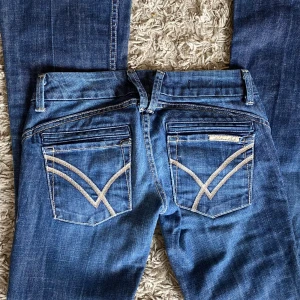 Blå bootcut jeans från Envoy - Säljer ett par blå bootcut jeans från Envoy med snygga broderade detaljer på bakfickorna. Jeansen har låg midja, klassisk femficksdesign och kontrastsömmar. Materialet är mjukt jeans med lite stretch för skön passform. Perfekt för dig som gillar retrovibe och markerade fickor.