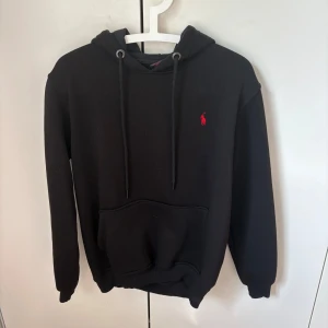 Svart hoodie från Polo Ralph Lauren - Svart hoodie från Polo Ralph Lauren i storlek S. Klassisk design med huva och snörning, stor magficka och rött broderat logomärke på bröstet. skön bomull kvalitet. Passar för både kille och tjej