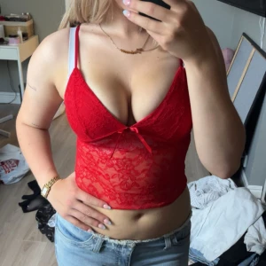 Röd spetsig topp - Säljer en röd topp med spets och liten rosett framtill. Toppen är ärmlös och har en djup v-ringning som ger en snygg siluett. Perfekt för dig som vill sticka ut med en färgstark och trendig look.