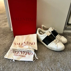 Valentino Garavani Open sneakers vit/svart - Säljer ett par snygga Valentino Garavani Open sneakers i vitt med bred svart rem över mitten. Skorna har rund tå, platt sula och klassisk snörning. Tillverkade i skinn och kommer med originalpåse och låda. 