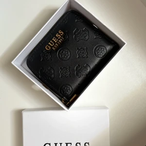 Svart plånbok från Guess med mönster - Snygg svart plånbok från Guess med präglade logomönster och guldfärgad text på framsidan. Plånboken har dragkedja runtom och flera fack för kort och mynt. Tillverkad i skinn och levereras i originalask. Perfekt storlek för att ha med sig överallt.