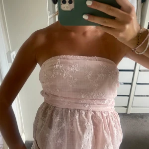 Ljusrosa bandeau tubtopp med broderade blommor - Superfin ljusrosa bandeau tubtopp med broderade vita blommor och lätt volang nedtill. Toppen är axelbandslös. 