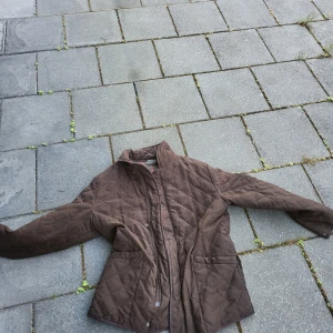 Brun quiltad jacka från Falcon - Quiltad brun jacka från Falcon med klassisk krage och långa ärmar. Jackan har tryckknappar och dragkedja framtill, samt två fickor. Insidan har ett rutigt foder i rött och beige. Perfekt för höst och vinter när du vill ha en chill och snygg look.