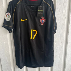 Portugal C. Ronaldo svart fotbollströja - Snygg svart Portugal fotbollströja från Nike med gult nummer 17 och C. Ronaldo på ryggen. Tröjan har korta ärmar, gul Nike-logga och Portugal-emblem på bröstet. Materialet är lätt och andas, perfekt för match eller träning. EM 2006 patch på ärmen.