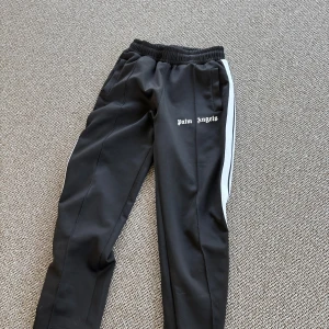 Palm Angels byxor - Svarta trackpants från Palm Angels med vita sidoremsor och logotyp på framsidan. Byxorna har elastisk midja och raka ben med sportig vibe. Tillverkade i Italien, materialet är troligtvis polyester. Perfekta för en streetwear-look., Mycket bra skick.