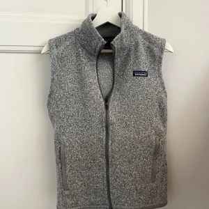 Grå fleeceväst från Patagonia - Säljer en gråmelerad fleeceväst från Patagonia i storlek S. Västen har hög krage, hel dragkedja framtill och två sidofickor med grå kant. Perfekt för lager-på-lager och chill utomhus. Patagonia-logga på bröstet.