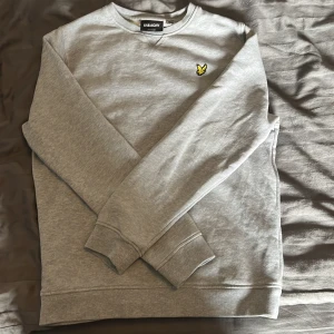 Grå sweatshirt från Lyle & Scott - Snygg grå sweatshirt från Lyle & Scott med klassisk rund hals och långärmad design. Tröjan har det ikoniska gula fågelmärket broderat på bröstet och är tillverkad i mjuk bomull för skön komfort. Perfekt för chill dagar och enkel att matcha med jeans eller joggers. Storlek 10-11 