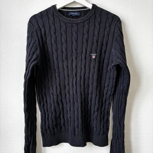 Mörkblå kabelstickad tröja från GANT - Mörkblå kabelstickad tröja från GANT med rund hals och diskret logga på bröstet. Tröjan är tillverkad i 100% bomull och har ribbade muddar vid ärmslut och nederkant. Perfekt för dig som gillar klassisk och stilren look.