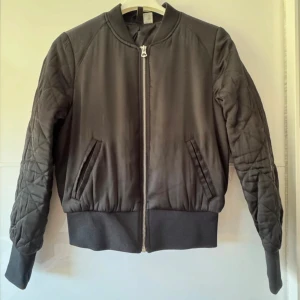 Svart bomberjacka från H&M Divided - Snygg svart bomberjacka från H&M Divided i storlek 36. Jackan har quiltade ärmar, ribbade muddar och dragkedja framtill. Tillverkad i 100% polyester, med två snedställda fickor och klassisk bomberkrage. Perfekt för dig som gillar streetstyle. Små skavanker på baksidan av jackan, se sista bilden.