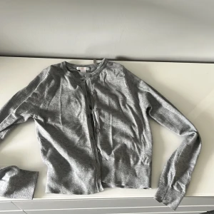 Grå kofta från Gina Tricot - Snygg grå kofta från Gina Tricot i storlek 158/164. Den har rund hals, långa ärmar och knappar framtill. Materialet känns mjukt och lätt, perfekt för lager-på-lager. Enkel och stilren design som funkar till många olika outfits.