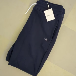 Mörkblå mjukisbyxor från GANT 146/152 - Snygga mörkblå mjukisbyxor från GANT med vit snörning i midjan och diskret logga på benet. Helt nya, tagg kvar. Byxorna har ribbad mudd vid midja och benslut för extra komfort. Perfekta för chill eller häng med kompisar.