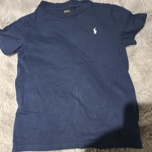 Marinblå t-shirt från Polo Ralph Lauren - Snygg marinblå t-shirt från Polo Ralph Lauren med klassisk vit logga broderad på bröstet. T-shirten har rund hals och korta ärmar, tillverkad i mjuk bomull som är skön mot huden. Perfekt för dig som gillar stilrena och tidlösa plagg.