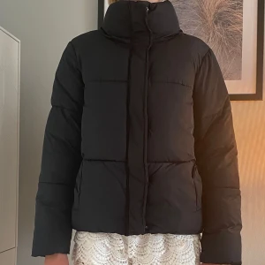 Svart pufferjacka med hög krage - Säljer en svart pufferjacka med hög krage och raka, quiltade paneler. Jackan har långärmade ärmar och stängs med dragkedja framtill. Perfekt för kalla dagar och har en clean, stilren look som funkar till de flesta outfits.