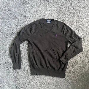 Ralph Lauren tröja  - Svart v-ringad tröja från Polo Ralph Lauren i slim fit med lila broderad logga på bröstet. Tillverkad i mjuk pima bomull, har långa ärmar och ribbade muddar vid ärmslut och nederkant. Perfekt för dig som gillar stilrena och klassiska plagg.