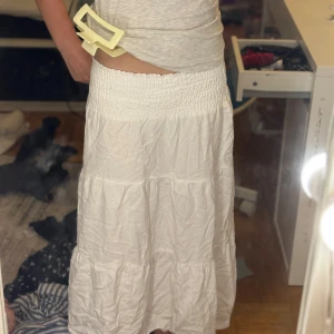 Vit långkjol med smock i midjan - Säljer en vit långkjol med bred smock i midjan som ger en skön och flexibel passform. Kjolen har volanger och är tillverkad i ett lätt, krinklat tyg som känns luftigt och somrigt. Perfekt för dig som gillar en chill och bohemisk stil. Stryka såklart innan köp!