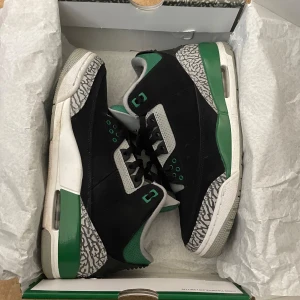 Jordan 3 Retro - Nike Air Jordan 3 Retro sneakers med svart mocka, gröna och grå detaljer. Skorna har snörning, perforerad grå plös med grönt Jumpman-logo och platt sula med synlig Air-enhet. Perfekt för dig som gillar streetwear. Äkta såklart.