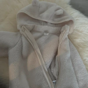 Beige teddyjacka med öron på huvan - Supermysig beige teddyjacka med huva som har söta öron. Jackan är fluffig och har en dragkedja framtill. Perfekt för att hålla sig varm och se gullig ut. Materialet är mjukt fleece och modellen är loose fit.