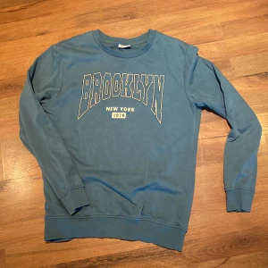 Blå Brooklyn tröja från Kappahl - Snygg blå sweatshirt från Kappahl med stort BROOKLYN-tryck i vitt och beige på bröstet. Tröjan har rund hals, långa ärmar och ribbade muddar. Materialet är mjuk bomull blandat med polyester, perfekt för chill dagar. Passar dig som gillar streetstyle och enkel design.